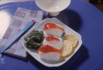 Photograph shows a plate with sushi on rice, slices of lemon, and some green food item, probably seaweed, on a mat with a bowl, napkin, and pair of chopsticks, 1970s. (Photo by Hulton Archive/Getty Images)